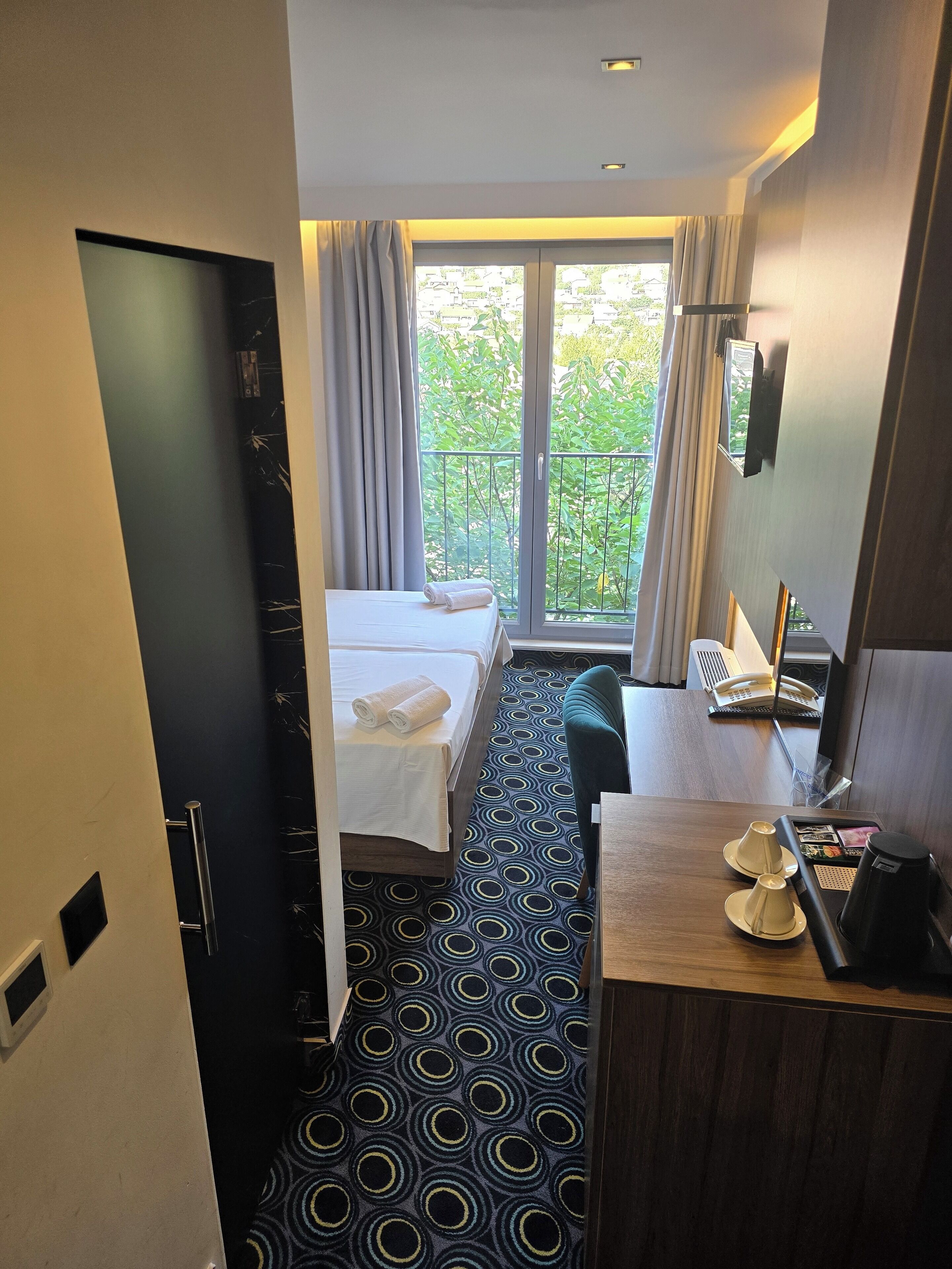 A hotel room with a bed, a desk, a chair, a coffee maker, and a view of greenery outside.