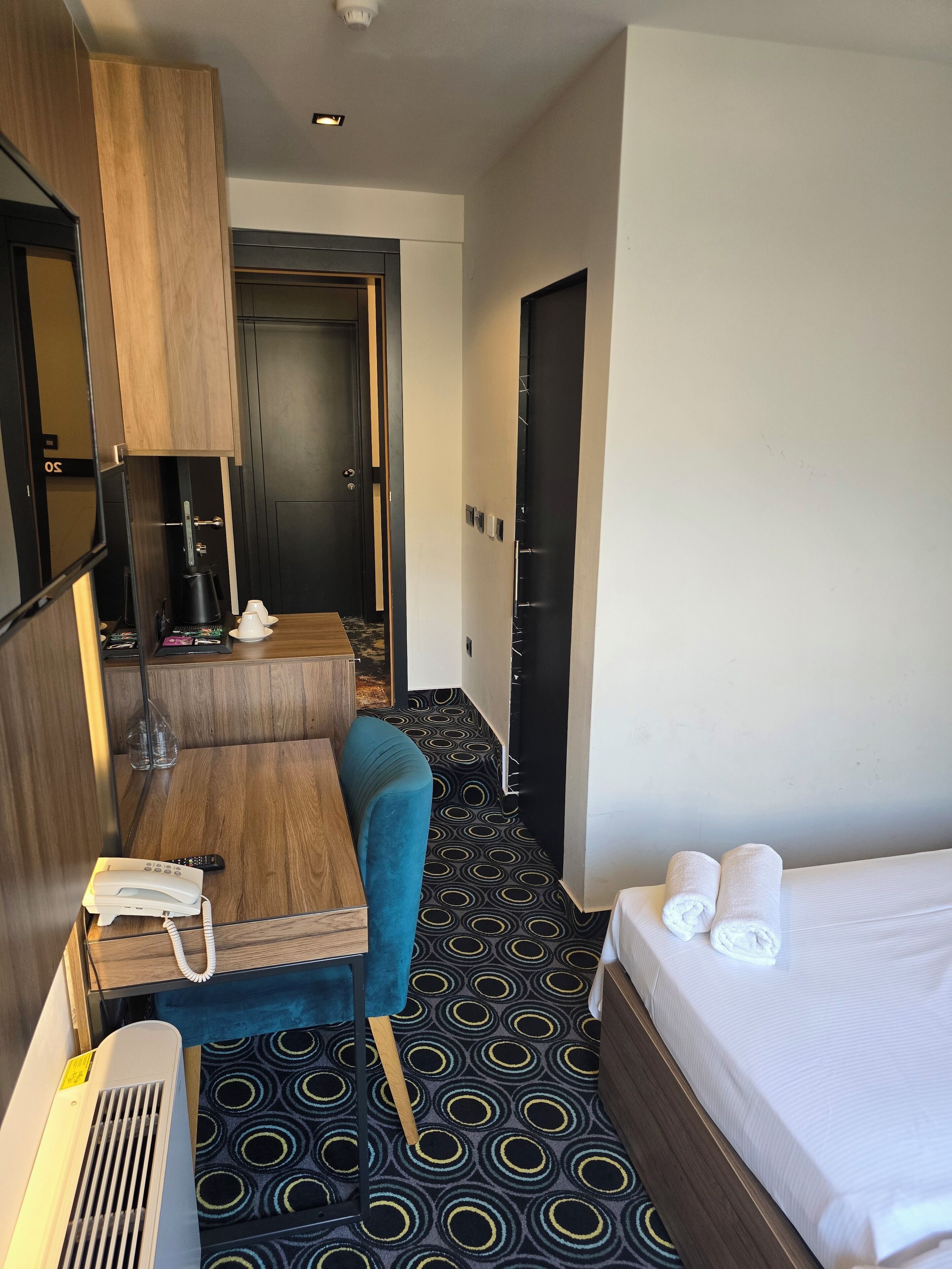 A hotel room with a bed, a desk with a telephone, a chair, a small table with a coffee pot, and a patterned carpet.