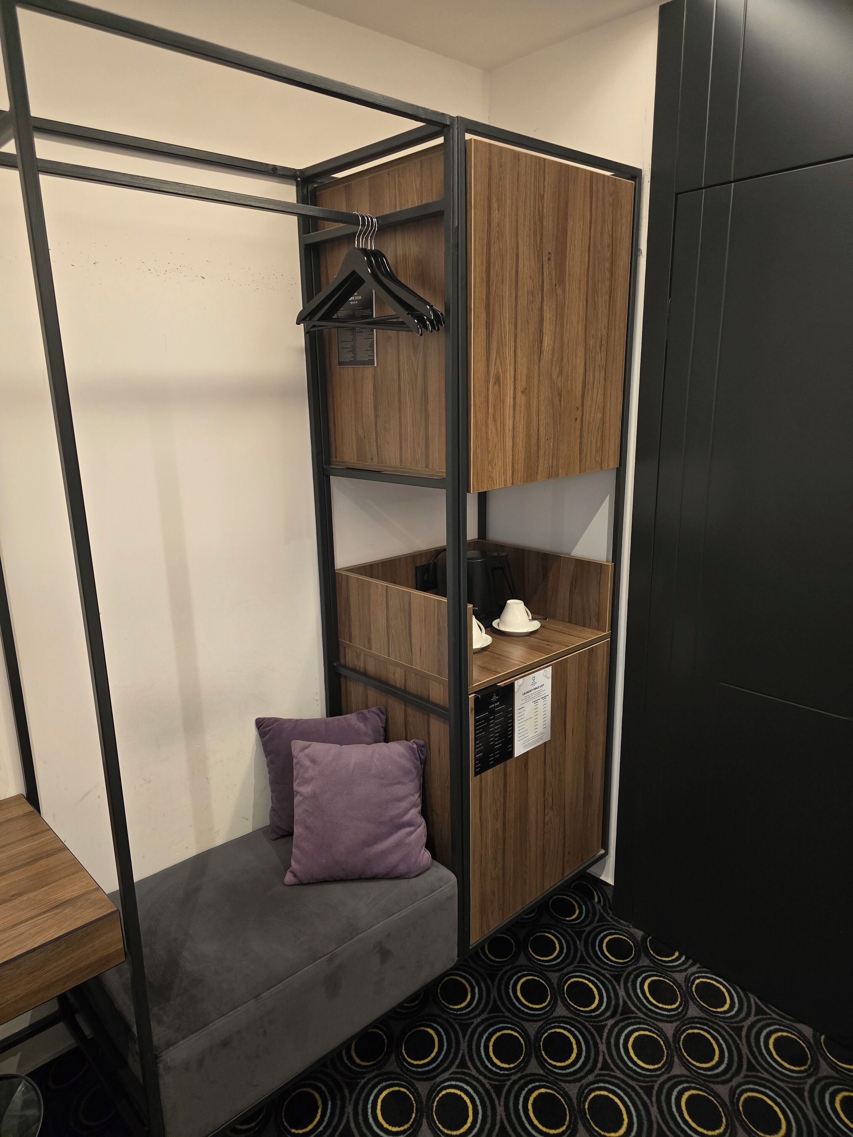 Quarto compacto com uma cama, uma mesinha, um travesseiro roxo, um cabideiro e uma porta preta.