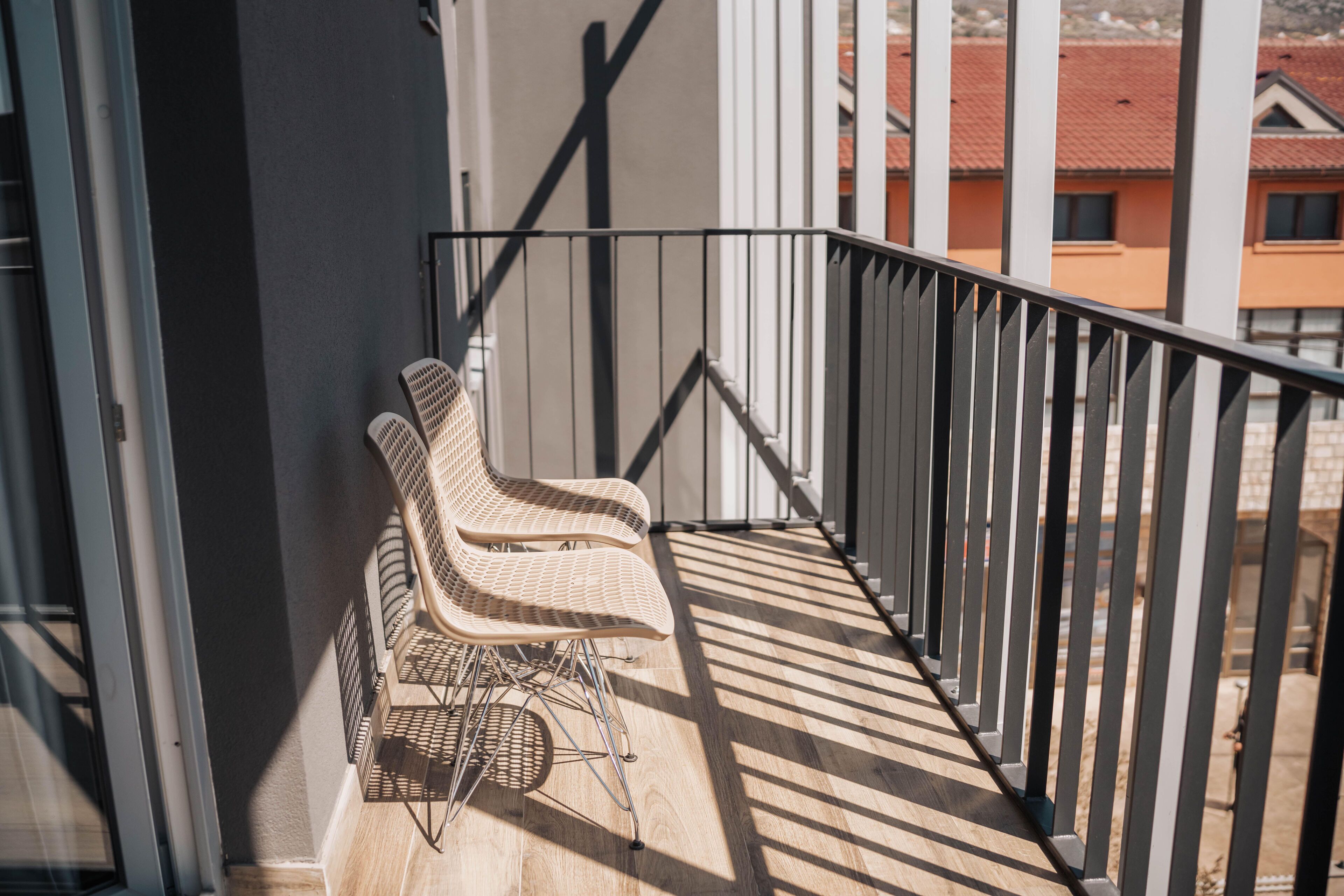 Uma varanda com duas cadeiras de vime, grade de metal e vista para edifícios ao fundo.