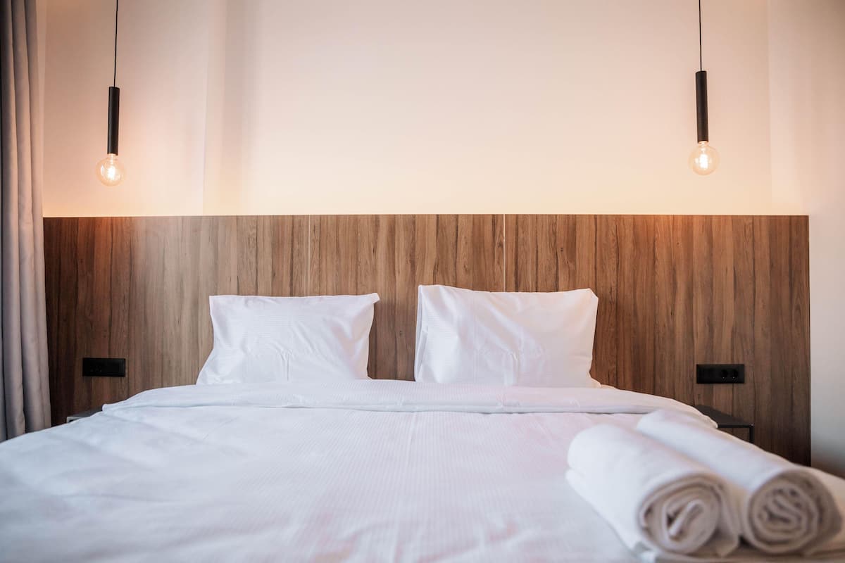 A neatly made bed with white linens and rolled towels, a wooden headboard, and two pendant lights.