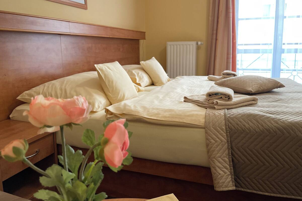 A hotel room with a bed, pillows, a bedside table, a vase with flowers, and a window with curtains.