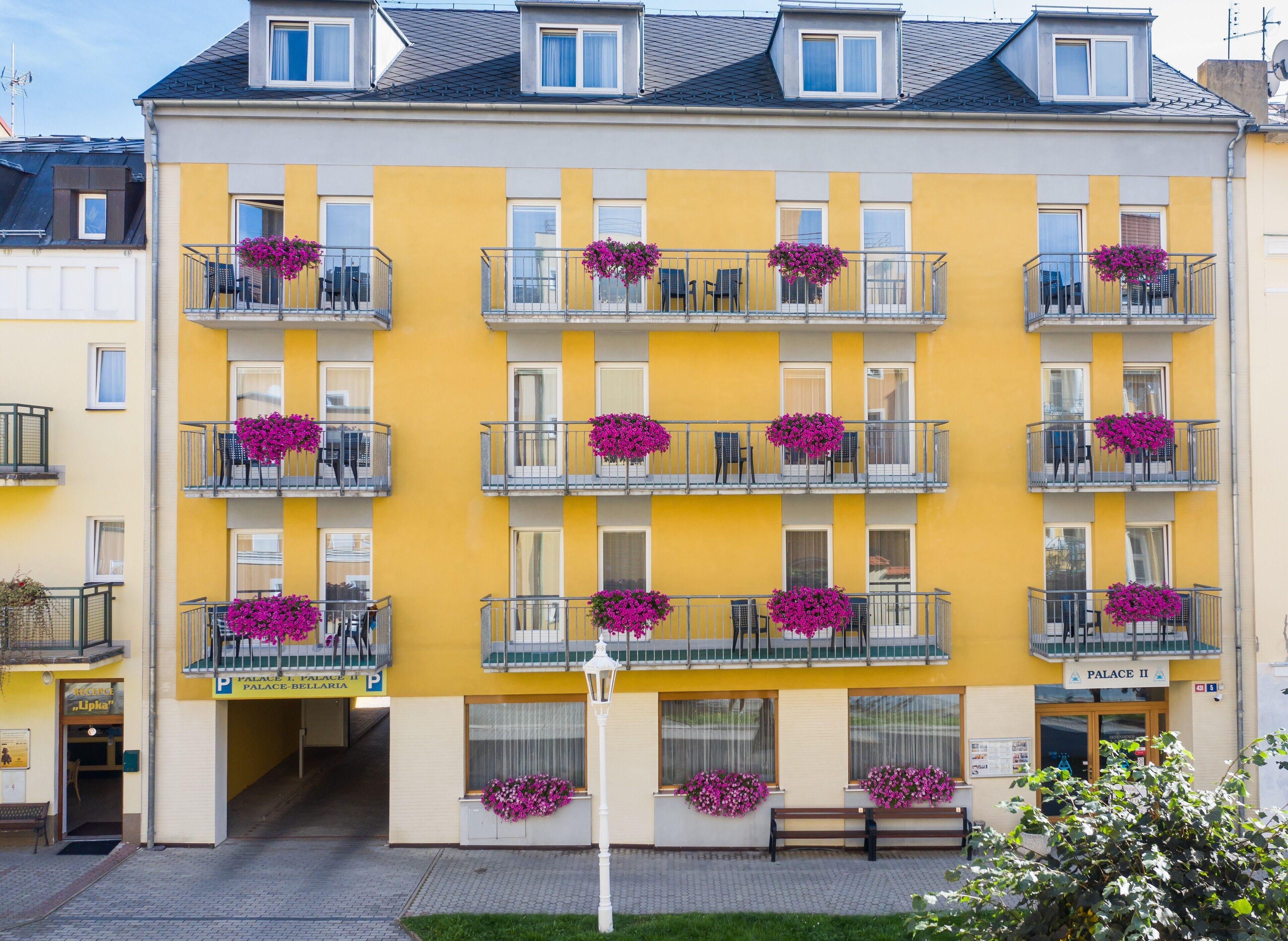 A multi-story building with balconies adorned with pink flowers, a sign for "Palace II," and a visible entrance.