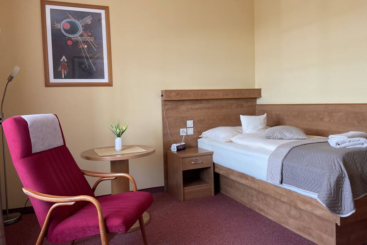 A hotel room with a single bed, a wooden headboard, a red armchair, a small table with a plant, and a framed abstract painting on the wall.