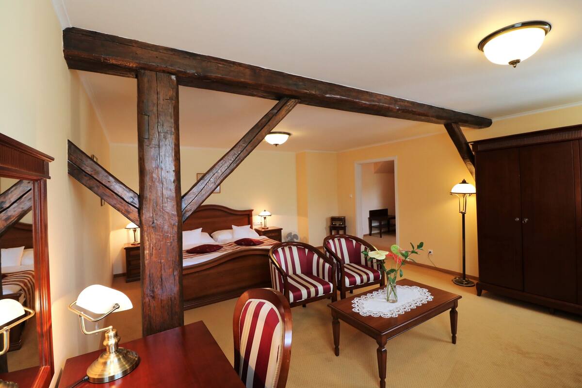 A hotel room with a wooden ceiling, a bed, two striped armchairs, a small table with a vase, and a nightstand with a lamp.