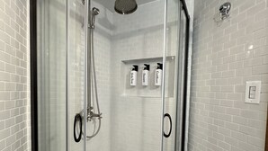 A glass-enclosed shower stall with a curved door, a handheld showerhead, and a wall-mounted faucet. There are three bottles of shampoo, conditioner, and body wash on a shelf above the shower. - The Walnut House - Right at OSU Greek Row and blocks from Theta Pond (Stillwater)