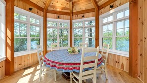 Una habitación con paneles de madera, una mesa redonda, sillas blancas y ventanas amplias con vistas a árboles y agua.