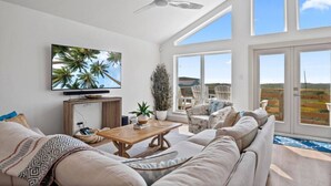 A spacious living room with a large TV, comfortable seating, a ceiling fan, and large windows offering a view of the outdoors. - Your Beach Retreat Awaits: Game Room, Community Pool & Easy Beach Access (Port Aransas)