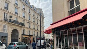 Straßenansicht mit einem Café, das Sitzgelegenheiten im Freien, eine rote Markise und ein mehrstöckiges Gebäude mit Balkonen im Hintergrund bietet.