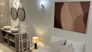 Ein modernes Hotelzimmer mit Bett, Nachttisch, Lampe, Spiegel und einem Badezimmer mit Waschbecken und Handtuchhalter.