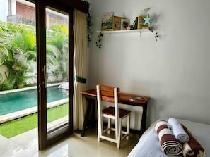 Une chambre d’hôtel avec vue sur une piscine, un bureau en bois avec une chaise, un lit avec des serviettes pliées, et une étagère avec des livres et une décoration en forme d’étoile de mer.