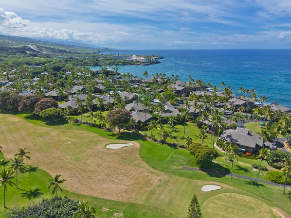 Vista aérea de un complejo turístico costero con varios edificios, palmeras y un campo de golf.