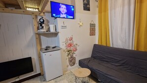 Een kleine, gezellige kamer met een grijze bank, een witte minibar, een aan de muur bevestigde televisie en een geel gordijn.
