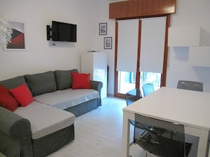 A living room with a sofa, a dining table, and chairs. There is a TV mounted on the wall, a window with blinds, and a door leading outside.