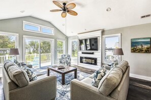 A spacious living room with a ceiling fan, large windows, a fireplace, and a television. - Sound View! Dogs OK! Pool&Spa, Sound&Beach Access (Corolla)