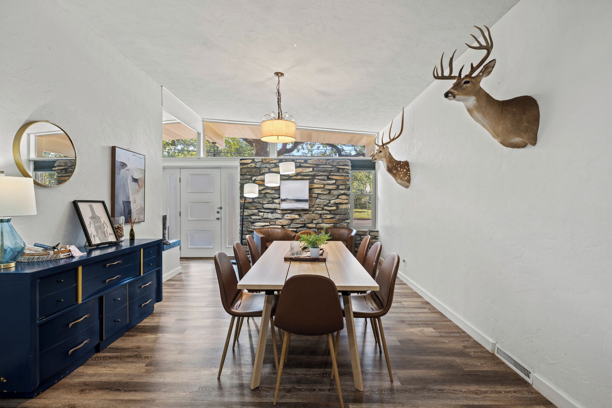 Ein Essbereich mit einem langen Holztisch, braunen Stühlen, einem aufgehängten Hirschkopf an der Wand und einer Steinwand im Hintergrund.