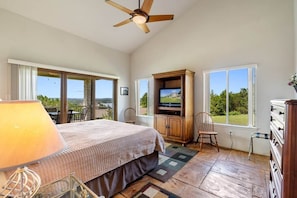 A bedroom with a bed, a television, a wooden cabinet, a chair, and a view of the outdoors.
