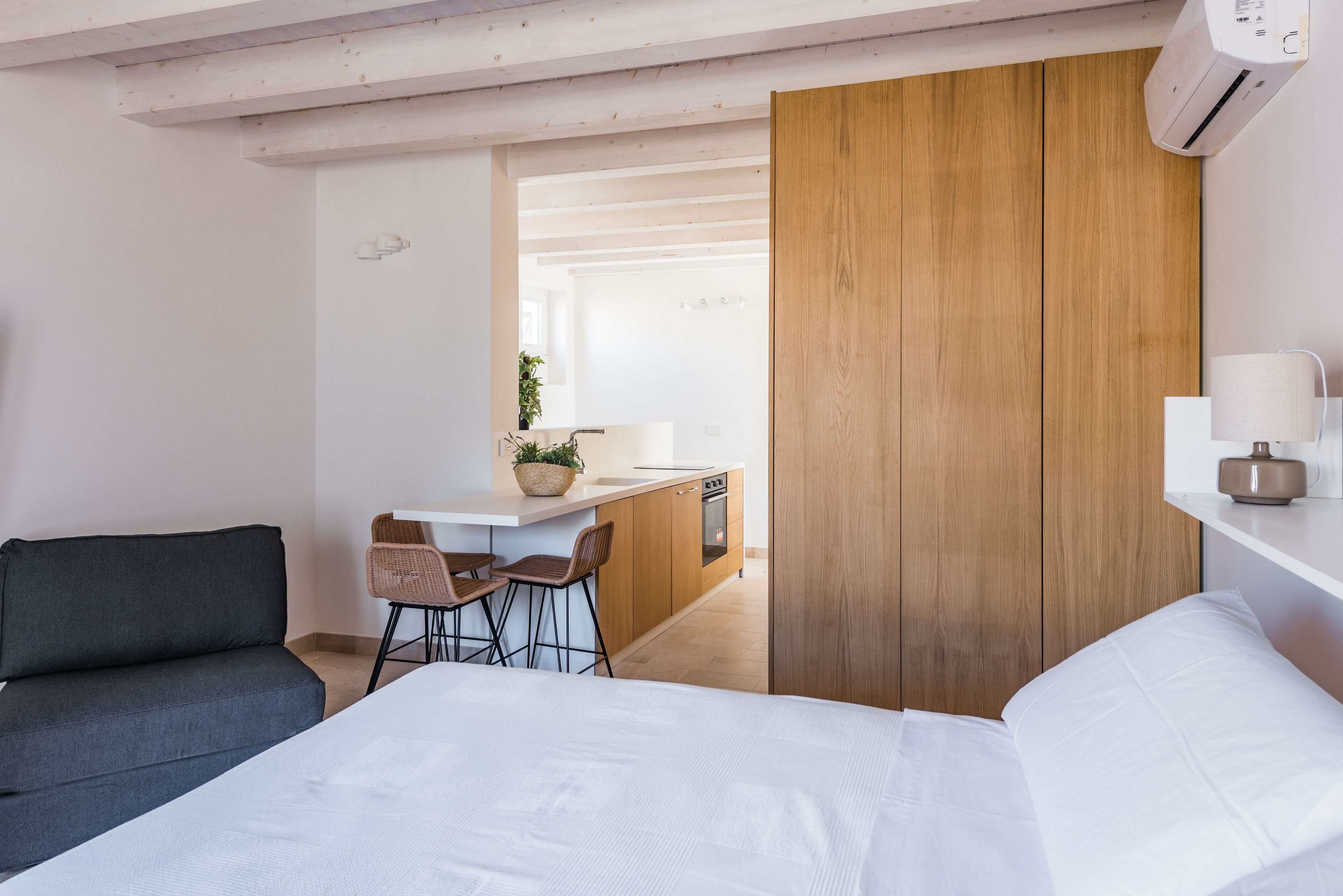 A modern hotel room with a bed, a sofa, a small kitchen area, and a wooden wardrobe.