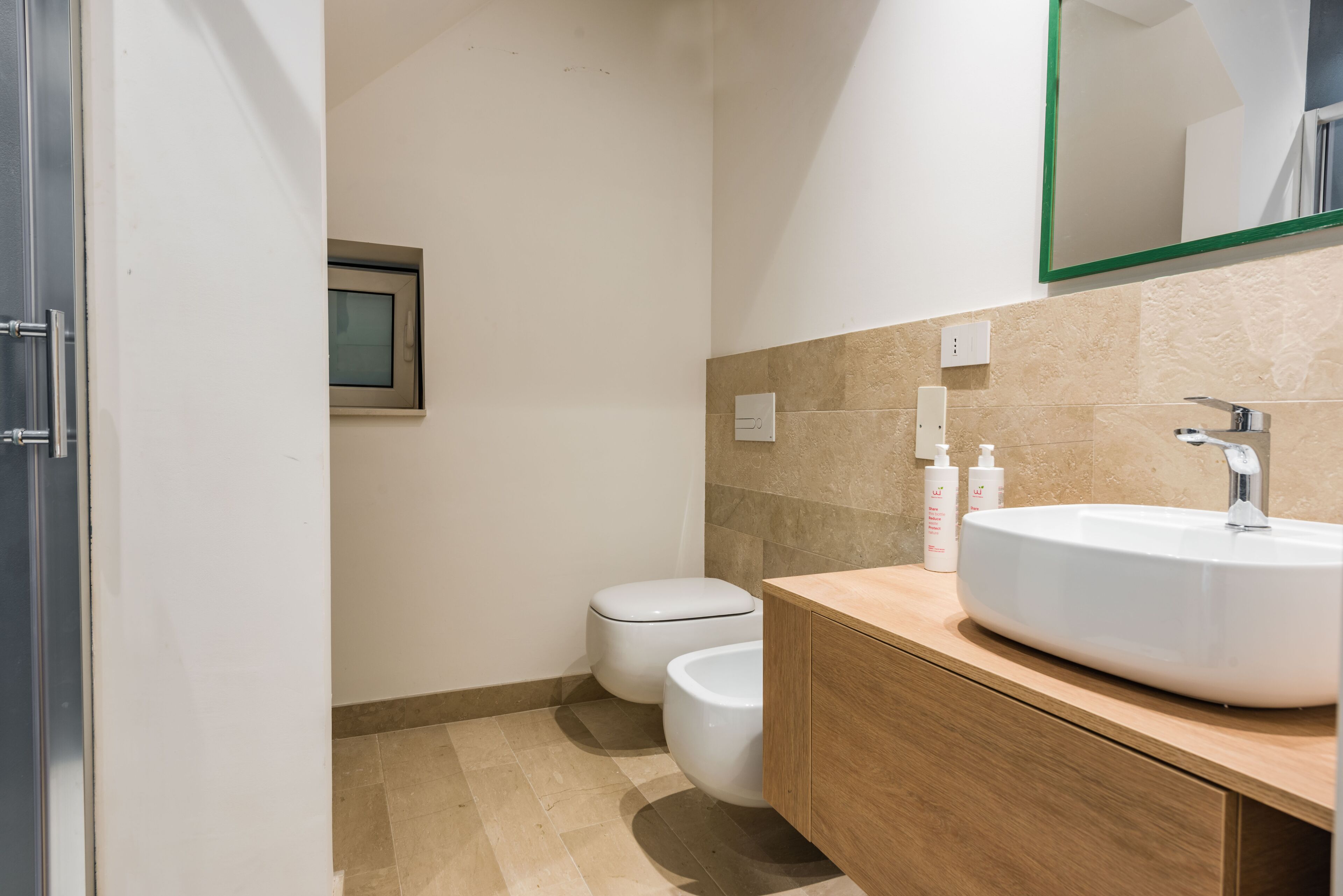 A modern bathroom with a white sink, wooden vanity, beige tiles, a toilet, and a mirror with a green frame.
