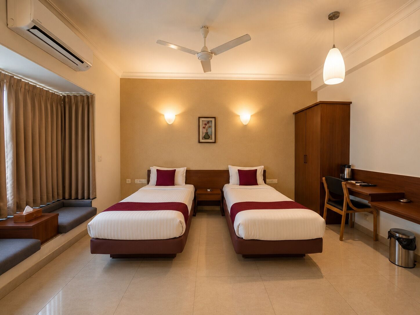 A hotel room with two beds, a desk, a chair, an air conditioning unit, a ceiling fan, and a picture on the wall.