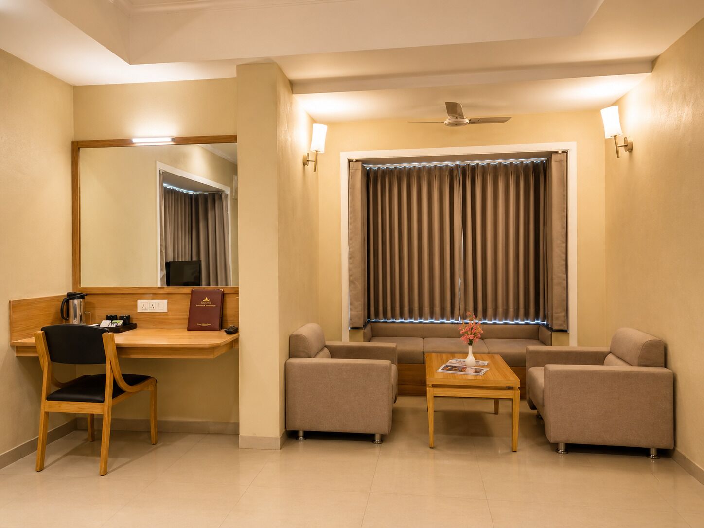A hotel room with a desk, two armchairs, a sofa, a small table, a mirror, a ceiling fan, and a window with curtains.