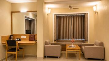 A hotel room with a desk, two armchairs, a sofa, a small table, a mirror, a ceiling fan, and a window with curtains.