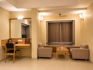 Une chambre d’hôtel équipée d’un bureau, de deux fauteuils, d’un canapé, d’une petite table, d’un miroir, d’un ventilateur de plafond et d’une fenêtre avec des rideaux.