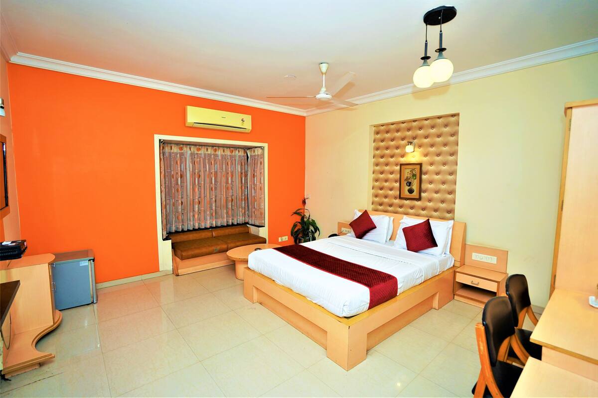 A hotel room with a bed, a desk, a chair, a small table, a ceiling fan, and a window with curtains.
