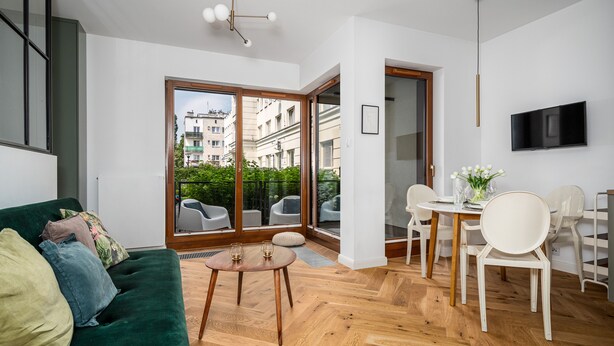 A modern living room with a green sofa, a wooden dining table with chairs, and a balcony with a view of buildings. - Apartamenty Sun & Snow Rezydencja Łazienki Park (Warsaw)