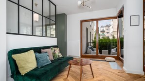 A modern living room with a green sofa, a small wooden table, and a sliding glass door leading to an outdoor area with chairs and a view of buildings. - Apartamenty Sun & Snow Rezydencja Łazienki Park (Warsaw)
