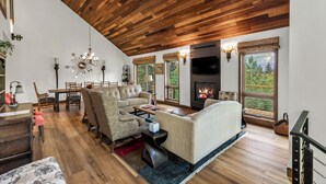 A spacious living room with a high wooden ceiling, a fireplace, and large windows offering a view of the outdoors.