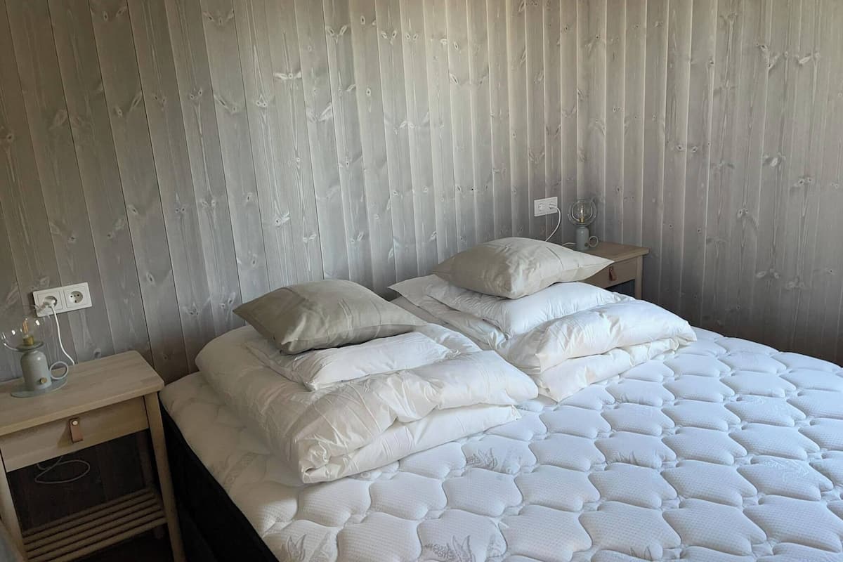 En pænt redt seng med hvide sengetøj og puder, et natbord med en lampe, og en stikkontakt på væggen.