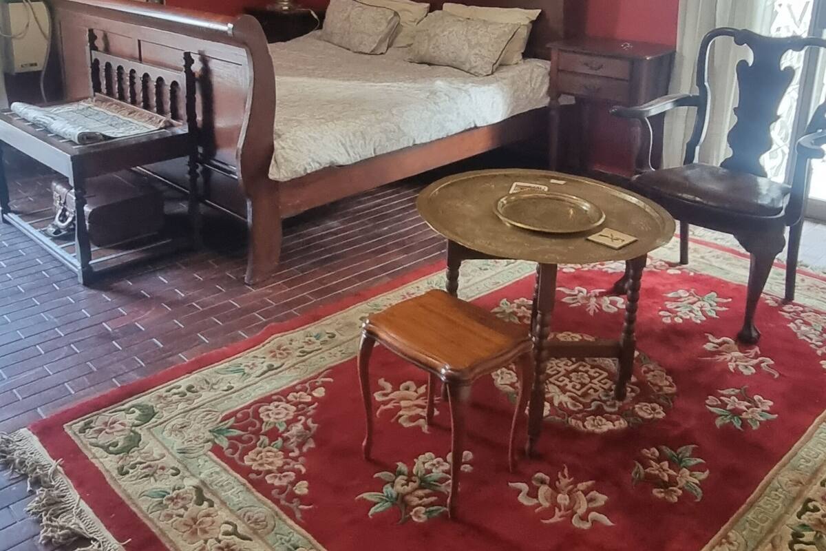 A room with a bed, a wooden chair, a small table, and a red carpet with floral patterns.