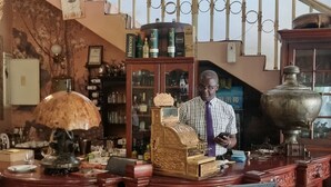 A bar area with ornate wooden furniture, a cash register, and a man standing behind the counter. - The Gallery Boutique Hotel (Harare)