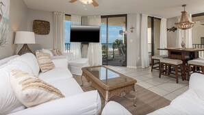 A spacious living room with a white sofa, a glass coffee table, and a dining area with a wooden table and chairs. A large TV is mounted on the wall, and a balcony with a view of the beach is visible. - 2ND Floor Condo at Phoenix V! (Orange Beach)