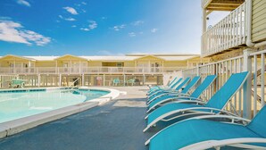 Zona de piscina con sillas reclinables azules y una piscina, rodeada por una valla blanca y edificios amarillos.