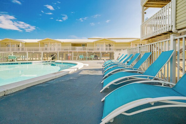 A pool area with blue lounge chairs and a pool, surrounded by a white fence and yellow buildings. - Home Away From Home at Moonraker 20 (Gulf Shores)