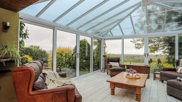 A conservatory with a glass roof, furnished with leather sofas and a wooden table, surrounded by greenery.
