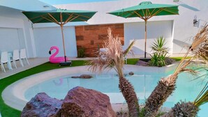 A swimming pool with rocks and palm trees, surrounded by a patio with chairs and umbrellas.
