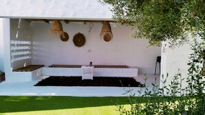 A modern outdoor seating area with a white bench and a small table, surrounded by greenery and a white wall with woven decorations.