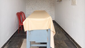 A narrow room with a bed, a clock on the wall, a red chair, and a wall-mounted electrical outlet. - JnJ Homestay (Chikkamagaluru)