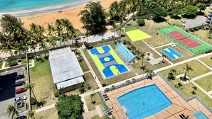 Vista aérea de uma área costeira com praia, palmeiras, quadra de tênis, quadra de basquete e piscina.