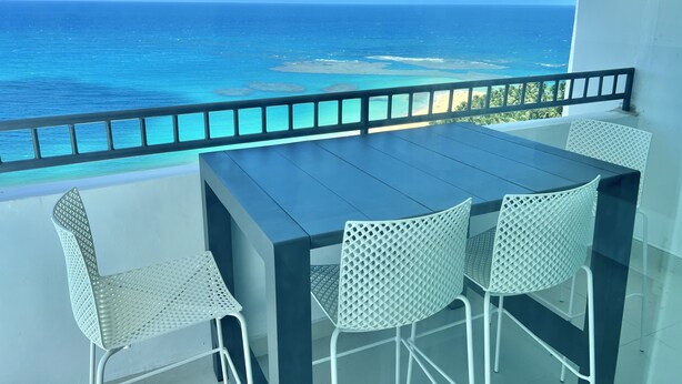 Varanda com mesa e cadeiras, com vista para um mar azul-turquesa e uma praia.