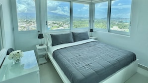 Quarto com cama, mesa de cabeceira, luminária e vista para uma paisagem através de grandes janelas.