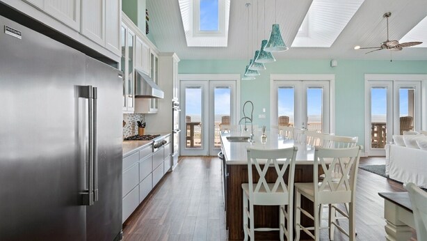 A modern kitchen with a stainless steel refrigerator, white cabinetry, and a large island with white chairs. There are skylights and a ceiling fan.