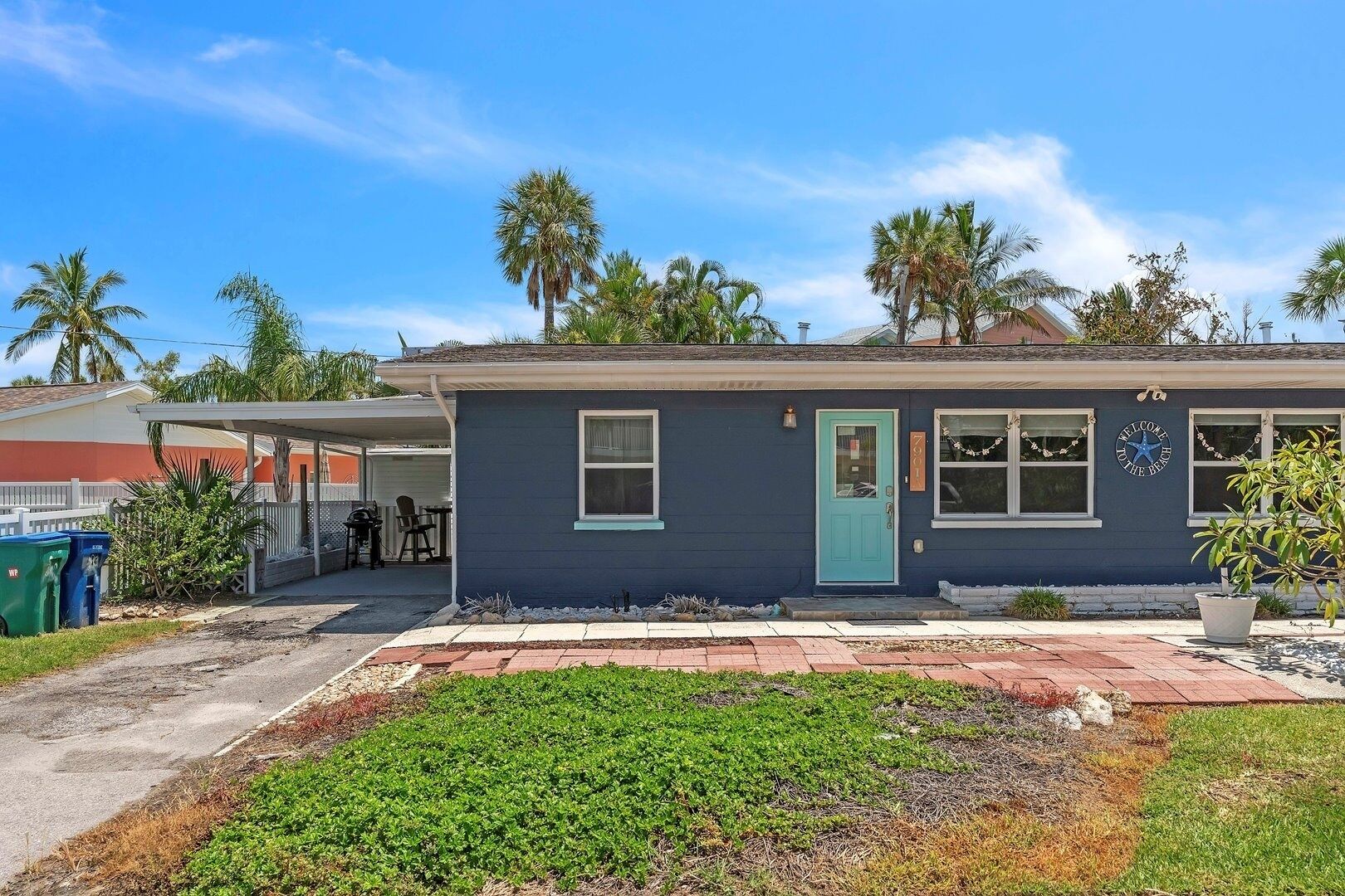 A single-story house with a dark blue exterior, a turquoise door, and a small porch. There is a sign on the front door that reads "WELCOME HOME."