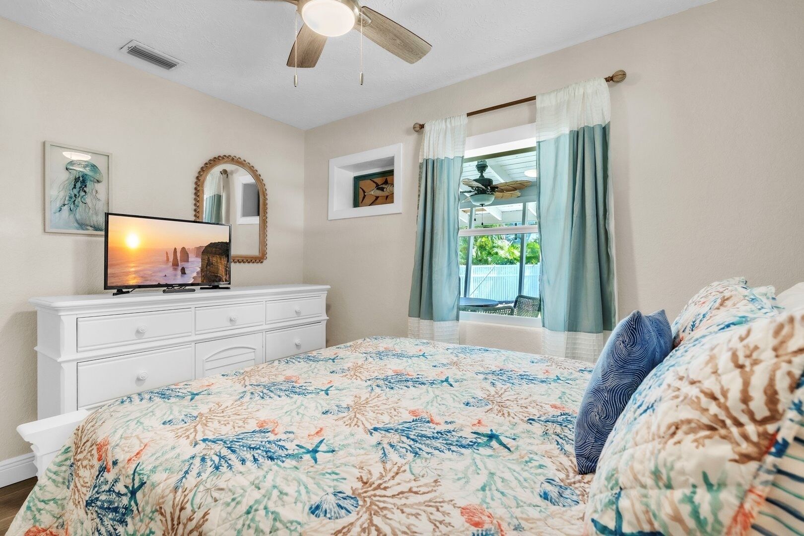 Une chambre à coucher comprenant un lit, une commode, un téléviseur, un ventilateur de plafond et une fenêtre avec des rideaux.