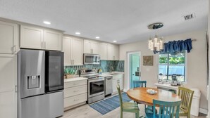 A modern kitchen with stainless steel appliances, white cabinets, and a dining area with a wooden table and chairs. - Seaduction Ami B 2 Bedroom Condo by RedAwning (Holmes Beach)