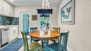 A dining area with a round wooden table, four chairs, a chandelier, and a kitchen with white cabinets and a blue backsplash. - Seaduction Ami B 2 Bedroom Condo by RedAwning (Holmes Beach)