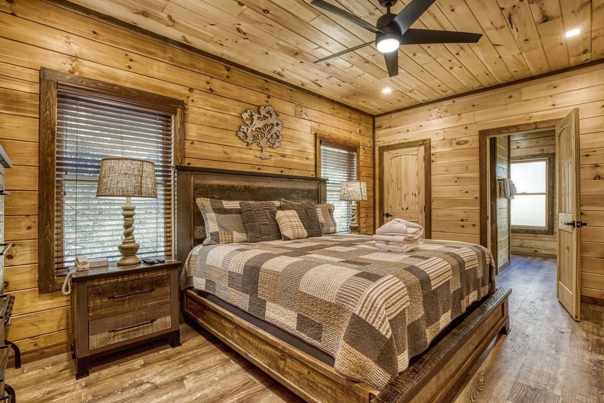 Dormitorio de cabaña de madera con cama, mesitas de noche, ventilador de techo y una puerta.